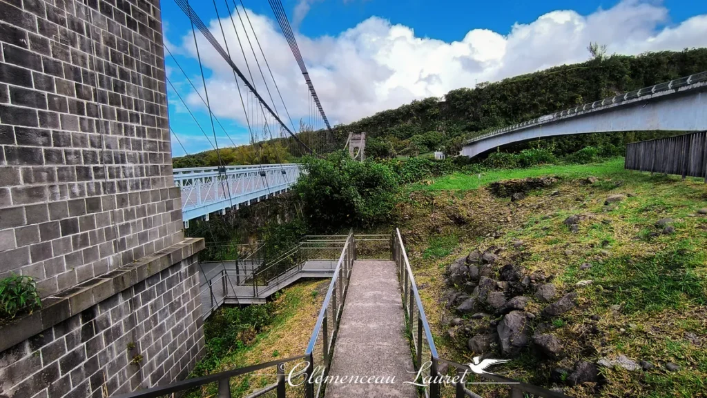 Vue plongeante depuis un escalier d'accès longeant la pile en pierre du pont suspendu. On aperçoit le tablier du pont historique sur la gauche, le pont routier en béton sur la droite et des chemins piétonniers aménagés sur des berges en herbe et rochers sombres. Le ciel est parsemé de grands nuages blancs. - Photo : Clémenceau Lauret (Licence CC BY-SA 4.0)