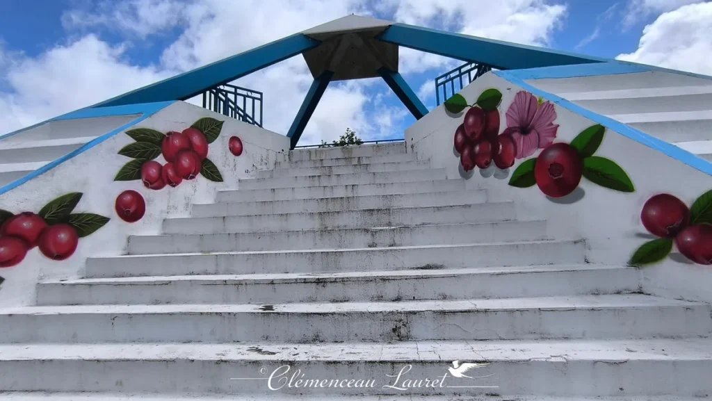 Escalier blanc en contre-plongée menant à la Pyramide Fleurie avec peintures de gouyaviers et hibiscus sous un ciel nuageux à La Plaine-des-Palmistes.
