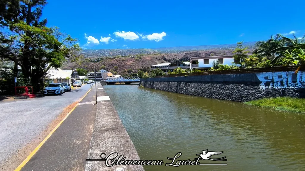 Vue panoramique en plongée du canal canalisé de la Ravine Grand Étang à Saint-Leu sous un ciel d'un bleu azur parsemé de quelques nuages blancs. Sur la gauche, la chaussée asphaltée bordée d'arbres tropicaux, de candélabres d'éclairage public et de véhicules garés longe un muret en béton et un trottoir. Au centre, le cours d'eau calme aux teintes sombres et verdâtres reflète la lumière du jour en s'étirant vers une passerelle piétonne métallique blanche au loin. À droite, un haut mur de soutènement en pierres basaltiques sombres, surmonté d'une paroi en béton marquée par des graffitis, borde la rive. En arrière-plan, les pentes rocheuses et arides des montagnes du littoral ouest réunionnais s'élèvent sous un soleil éclatant. Photo : Clémenceau Lauret à Saint-Leu (Licence CC BY-SA 4.0).