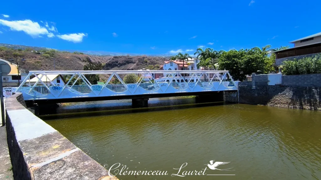 Vue rapprochée axiale d'une passerelle piétonne métallique blanche en treillis géométrique enjambant le canal de la Ravine Grand Étang à Saint-Leu. Le pont blanc, portant des inscriptions et graffitis sur ses montants, franchit les eaux paisibles du canal aux nuances verdâtres et ambrées. De part et d'autre, des murs de soutènement en pierre et des bâtiments résidentiels aux toitures rouges et grises s'intègrent au paysage sous un ciel bleu dégagé. En arrière-plan, les mornes et pentes sèches de la côte Ouest se découpent nettement. Photo : Clémenceau Lauret à Saint-Leu (Licence CC BY-SA 4.0).