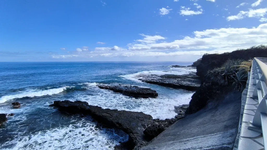 Vue en contre-bas depuis le muret de la piste sur la plateforme de basalte et l'écume des vagues sous un ciel bleu nuageux à Saint-Paul.