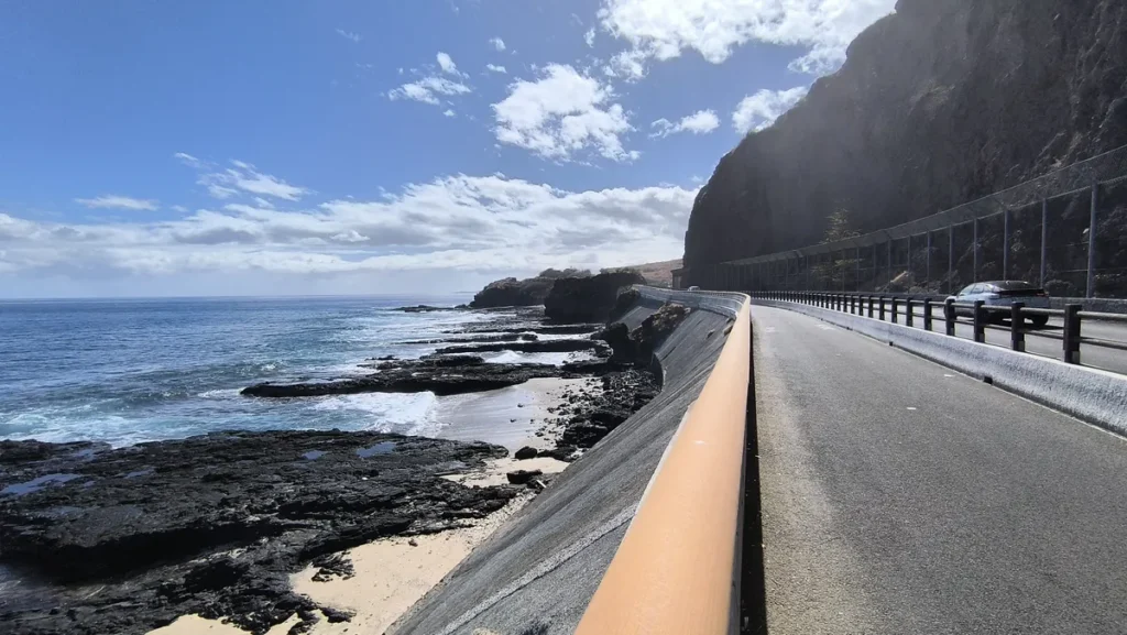 Voie goudronnée de la piste cyclable bordée d'une barrière orange longeant la falaise et la plage de basalte sous le soleil à Saint-Paul.