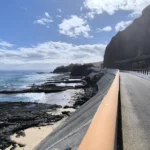 Voie goudronnée de la piste cyclable bordée d'une barrière orange longeant la falaise et la plage de basalte sous le soleil à Saint-Paul.