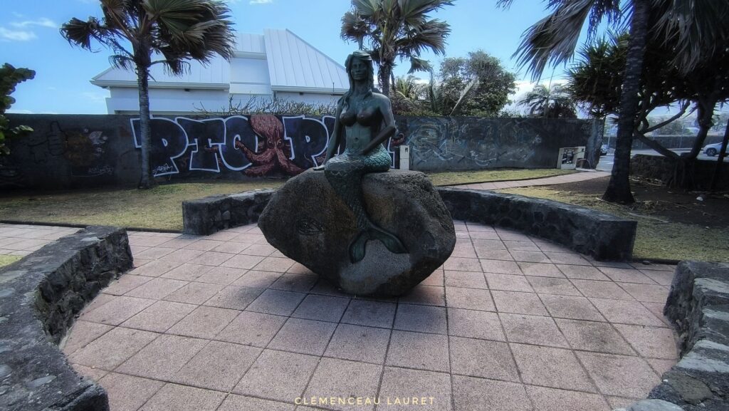 Vue de face de la statue en bronze de la sirène reposant sur un rocher au cœur d'une esplanade pavée à Saint-Pierre, entourée de palmiers et d'un mur décoré de graffitis - Photo : Clémenceau Lauret (Licence CC BY-SA 4.0)