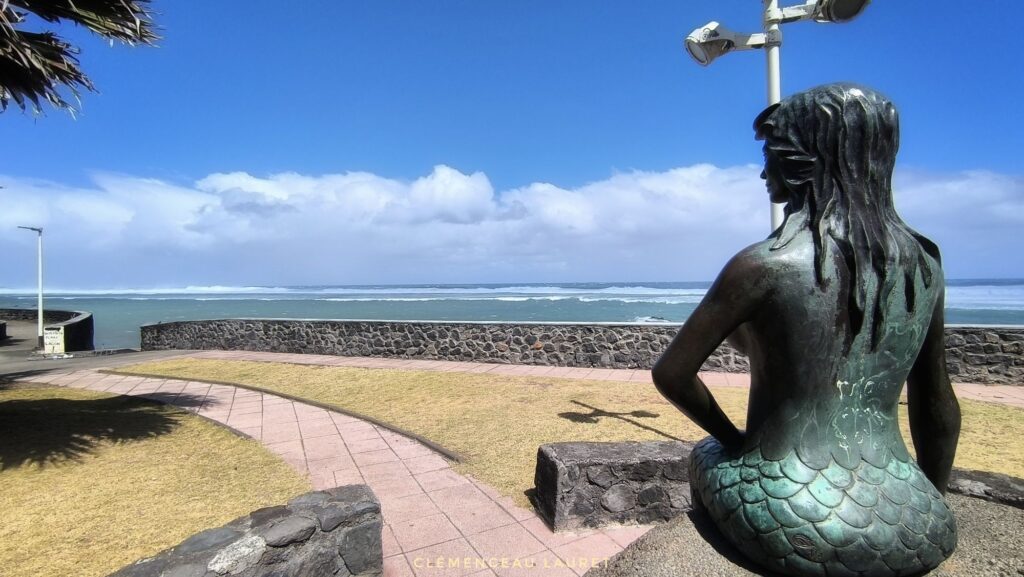 Statue en bronze d'une sirène assise de dos sur le front de mer de Saint-Pierre, contemplant l'océan Indien agité et la ligne d'écume blanche de la forte houle au large - Photo : Clémenceau Lauret (Licence CC BY-SA 4.0)