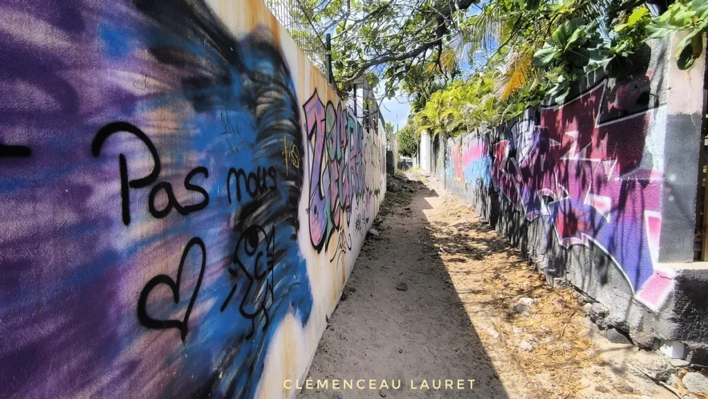 Murs tagués de cœurs et graffitis le long d'un sentier de sable blond à Saint-Gilles-les-Bains sous un ciel lumineux.