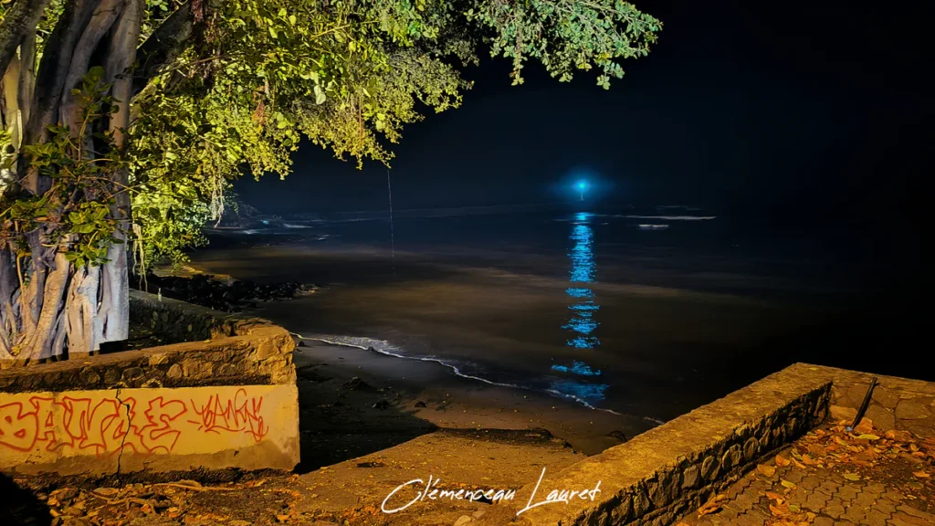 Plage nocturne de Terre-Sainte vue depuis un muret tagué, faisceau lumineux bleu-vert de la balise du port se reflétant sur la mer.