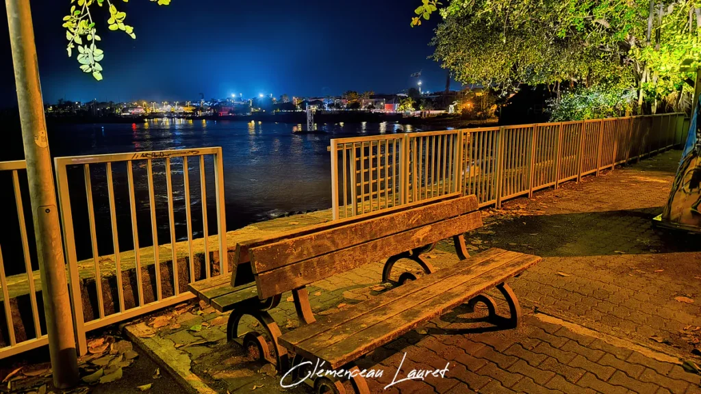 Banc en bois sur une promenade pavée la nuit à Terre-Sainte, garde-corps métallique et vue panoramique sur le port éclairé.